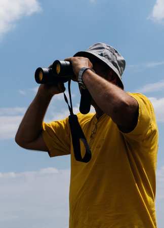 Man in a yellow T-shirt and panama looking through binocular in the distance against the sky. Aviation dispatcher or plane operator by the airfieldの写真素材