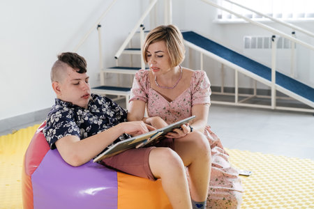 Children with disability reading book and communicating. Cerebral palsy boy talking with woman tale therapist. Rehabilitation center, mother with teenage person learning to speak, pronounce at homeの写真素材
