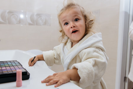 Toddler girl doing herself makeup with mamas cosmetics pretending grownup. Applying eyeshadows. Child in the bathrobe at homeの写真素材