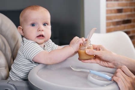 Mother giving food to baby boy, pure from spoon in highchair at home. Complementary infant feeding. Happy kid waiting for meal, learning to eat purred vegetables and fruitsの写真素材