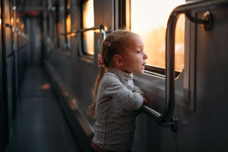 Toddler child looking though train window on sunset, bright sunlight, atmospheric travel by railway with kids. Girl happy exploring the way in evening. Exciting family tripの写真素材