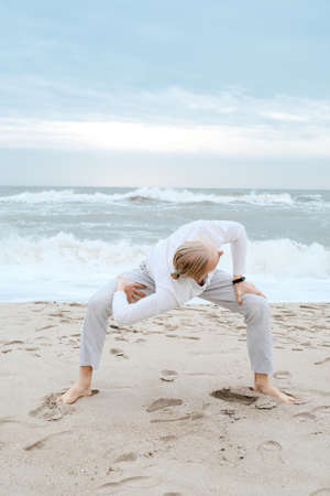 Man doing yoga on the beach in autumn over waves in the sea. Creamy, light sand and sky, Asanas and meditation, balance in life, self awareness and calmの写真素材