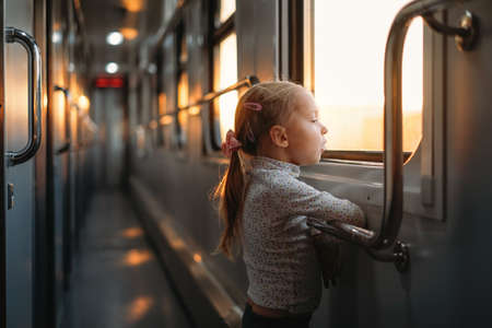Toddler child looking though train window on sunset, bright sunlight, atmospheric travel by railway with kids. Girl happy exploring the way in evening. Exciting family tripの写真素材