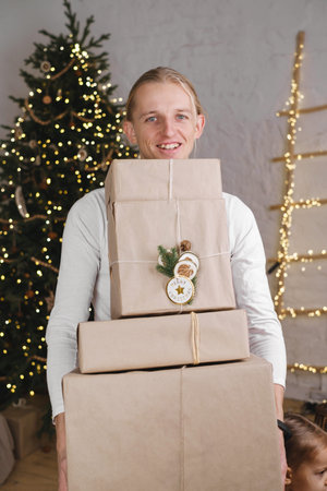 Happy man holding bed pile of Christmas gifts over decorated tree. Copy space, eco style, craft...