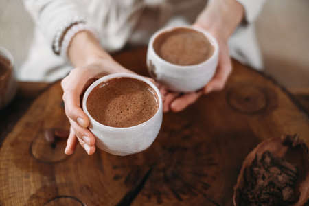 Hot handmade ceremonial cacao in white cups. Woman hands giving craft cocoa, top view on wooden table. Organic healthy chocolate drink prepared from beans, foam Giving cup on ceremony cozy atmosphereの写真素材