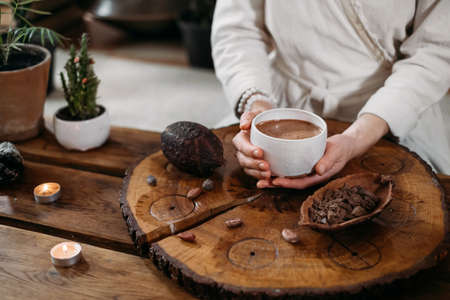 Hot handmade ceremonial cacao in white cup. Woman hands holding craft cocoa, top view on wooden table. Organic healthy chocolate drink prepared from beans, no sugar. Giving cup on ceremony, cozy cafeの写真素材