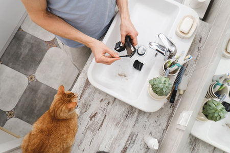 Man cleaning electronic shaving razor in sink, male self care and personal hygiene Top view. Hair in sink with cat. Long blonde hair, machine in handsの写真素材