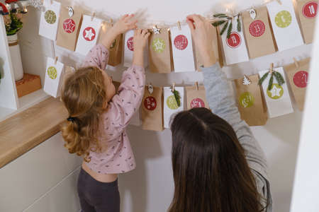 Toddler child gets new advent calendar task, waiting for Christmas. Festive mood for cute curly girl. Kid takes advent bag with dates from the wall at home.の写真素材
