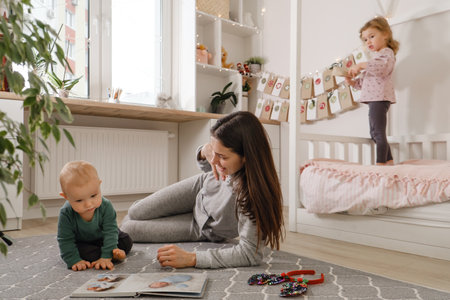 Mother spending time with two kids at home before Christmas. Looking photos in album, girl getting ready to open advent calendar tasks. Festive family time, happy children, siblings at homeの写真素材