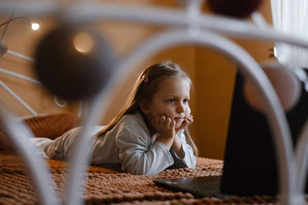Child watching cartoons on laptop. Serious kid with technology at home on bed spending time on-line. Education and entertainment for toddlers. Girl in cozy atmospheric room over the windowの写真素材
