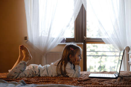 Child watching cartoons on laptop. Serious kid with technology at home on bed spending time on-line. Education and entertainment for toddlers. Girl in cozy atmospheric room over the windowの写真素材