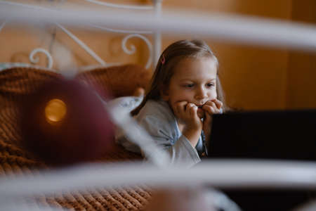 Child watching cartoons on laptop. Serious kid with technology at home on bed spending time on-line. Education and entertainment for toddlers. Girl in cozy atmospheric room over the windowの写真素材