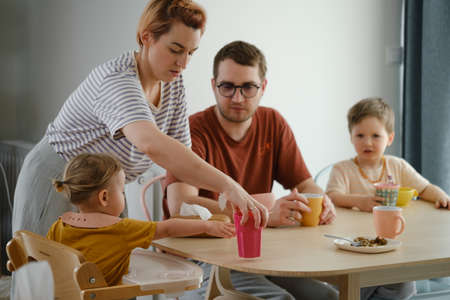 Parents with children having meal, breakfast by the table. Mother serving food to family at home. Eating together with kids. Girl in high chair eating, getting drinkの写真素材