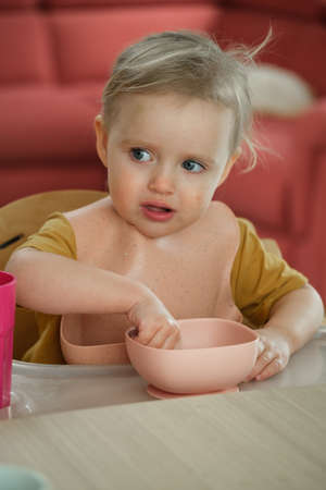 Toddler girl eating happily in high chair food by herself in the room. Child having meal. Happy kid having lunch. Little girl feeding herselfの写真素材