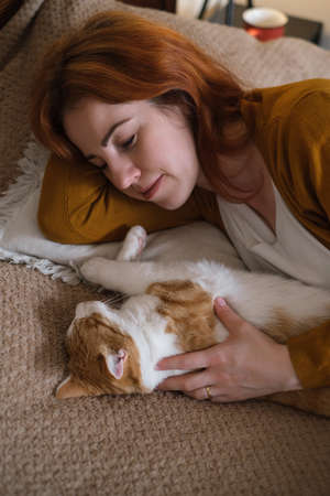 Middle age woman holding cute ginger cat. Female hugging her lovely short hair kitty in bedroom in the evening. Copy space. Adorable domestic pet concept.の写真素材