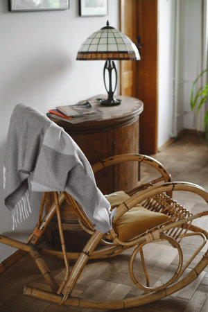 Old fashion wicker rocking chair with blanket in cozy house, lamp on wooden cabinet. Place for reading, no people, home plant near window. Comfortable vintage living room interiorの写真素材