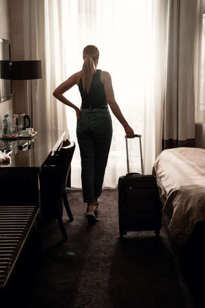 Woman traveler with luggage suitcase after checking Into a Hotel Room looking in the window. Silhouette of visitor in travel accommodation ready to have rest.の写真素材
