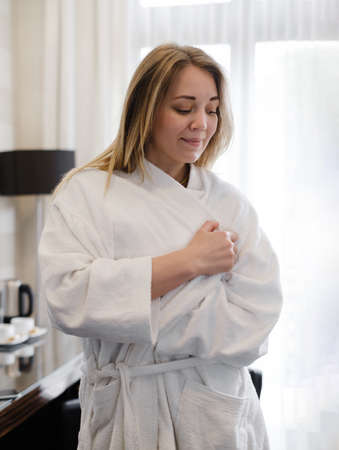 Woman wearing bathrobe in hotel room. Person in 20s in bedroom. Enjoying morning in cozy home atmosphere over the windowの写真素材