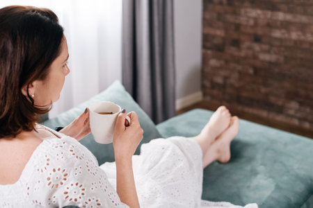 Woman drinking mug of coffee in cozy home atmosphere in the morning. Real middle age plus size female in white dress relaxing on the sofa, modern house. Sunny day with cup of tea, laying on couchの写真素材