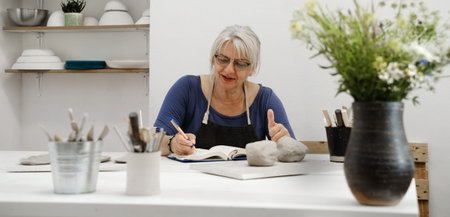 Banner Woman potter planning work at workshop, writing in notebook in light studio her creations....
