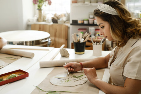 Woman decorating with flowers rolled clay, making ceramic plate in studio with floral pattern....