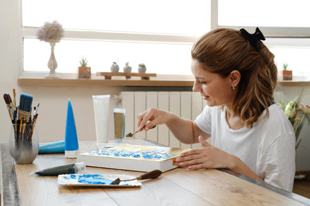 Woman artist painting picture at home with acrylic paints and spatula over the window. Adult hobby and free time creative activity. Female at her 40s enjoying art.の写真素材