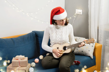 Woman celebrating Christmas alone at home, playing ukulele small guitar. Happy holidays lifestyle,...