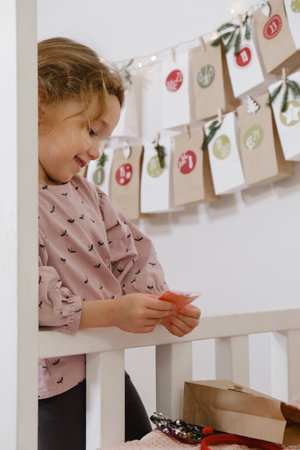 Toddler child gets new advent calendar task, waiting for Christmas. Festive mood for cute curly girl. Kid takes advent bag with dates from the wall at home.の写真素材