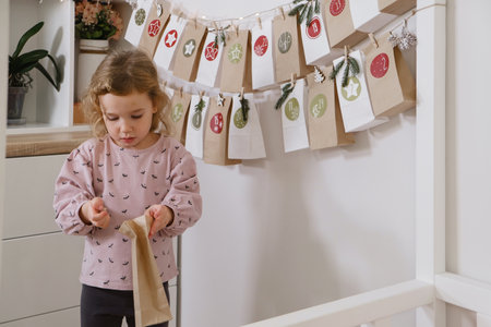 Toddler child gets new advent calendar task, waiting for Christmas. Festive mood for cute curly girl. Kid takes advent bag with dates from the wall at home.の写真素材