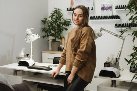 Nail service master. Portrait of professional young brunette sitting at modern nail salon workplace and smiling while waiting client. Beauty service Occupation conceptの写真素材