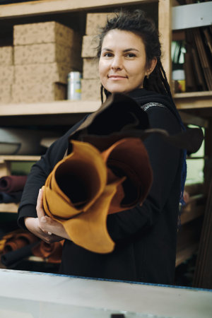 Tailor woman with fabric rolls. Pretty brunette designer looking camera while holding pile of leather rolls in workshop. Occupation concept. Small business owner. Female Textile Workerの写真素材