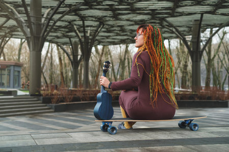 Funny free hipster Woman with dreadlocks sitting on longboard on the road playing guitar ukulele at...