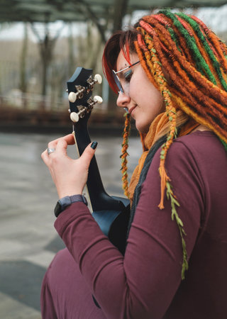 Funny free hipster Woman with dreadlocks sitting on longboard on the road playing guitar ukulele at her 30s. female with bright colorful hair enjoy outdoor active lifestyle, play musicの写真素材