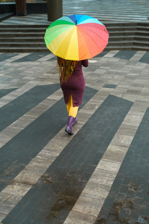 Rainbow umbrella on city street . Walking on a rainy spring day. Woman wearing rubber boots following the line. Bright colorsの写真素材