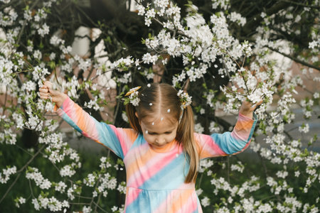 Nice little child in blooming garden exploring spring nature. Shaking tree making petals falling on her head. Fun in the garden.の写真素材