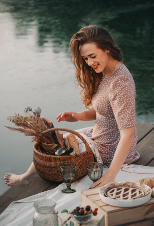 Woman on sunset on picnic by the water. Romantic evening portrait of female at her 30s in summer. Happy person at her free time festive decorated location on pier. Basket, pie and vineの写真素材