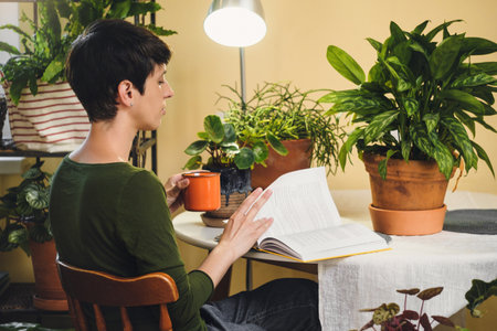 Woman at her reading place near houseplants. Green relax zone in the room with opened book and potted flowers. Female Enjoying peace and having rest at eco designed cozy house.の写真素材
