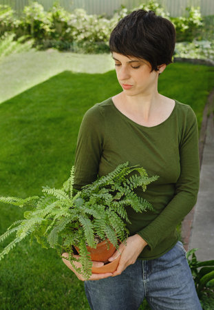 Woman holding houseplant over greengrass. Creative business concept. Confident female florist, owner of small business flower shop Holding potted fern outdoor. Enjoying job. Green on greenの写真素材