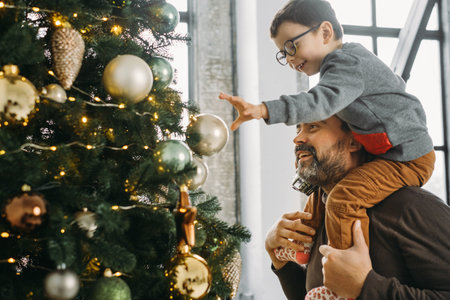 Father and child decorate Christmas tree at home. Son on dads shoulders putting bauble on tall tree.の写真素材