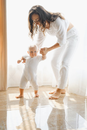 Mother helping to walk baby boy at home in a sunny room. Child learning to walk and keep balance...