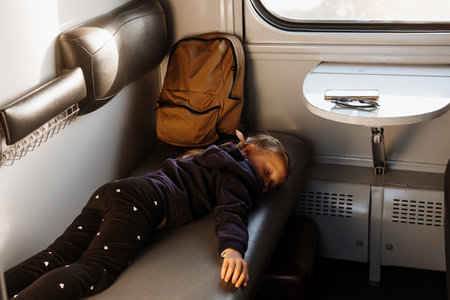 Tired child riding in train on sunset, bright sunlight, atmospheric travel by railway with kids. Passenger resting on the couch of slipping wagon. Long family tripの写真素材