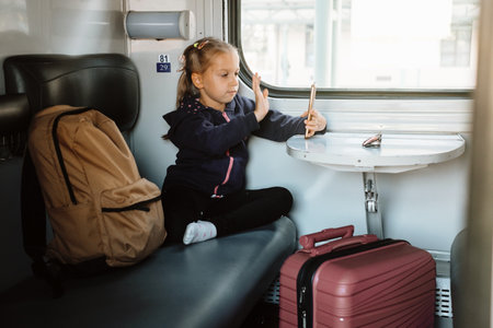 Child in train using smartphone. Kid communicating online while traveling. by railway. Girl sitting by the window with bags and gadget waving goodbyeの写真素材