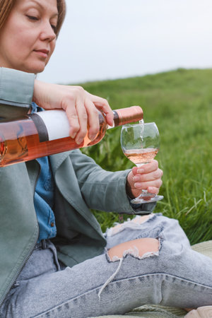 Woman uncork the bottle of rose wine on the picnic. Beautiful mature woman serve wine in glass outdoors on the grass. Woman at her 50s in the parkの写真素材