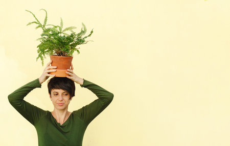 Woman holding houseplant on shoulder over yellow wall. Creative business concept. Confident florist, owner of small business flower shop Holding potted fern in workplace. Enjoying job. Bannerの写真素材