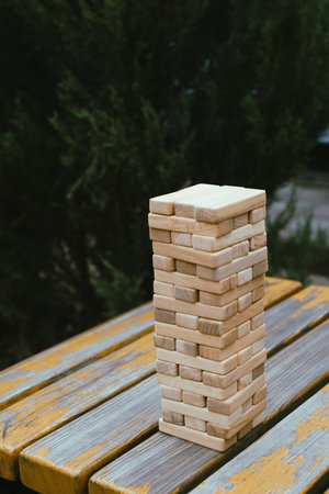 Person is stacking wooden blocks to build tower while seated at a table outdoors, concentrating on...