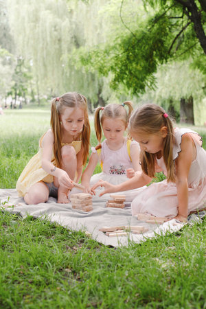 Three children gathered around wooden tower game, focused on stacking blocks, enjoying sunny day in the park. Friends playing tower game outdoorsの写真素材
