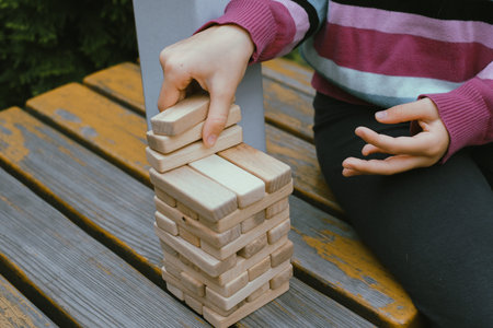 Person stacking wooden blocks, building tower while seated at a table outdoors, concentrating on...