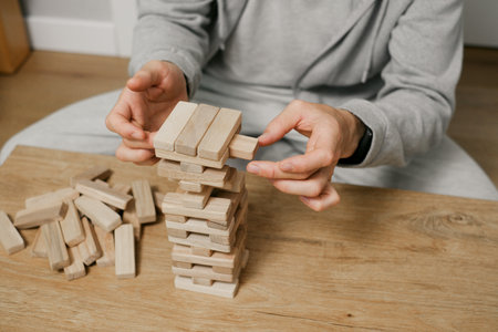 person stacks wooden blocks to construct tower on table in a room, Man playing table game for concentration at home. Digital detoxの写真素材