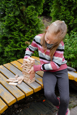 Preschool girl carefully balances wooden block with her fingers during game outdoors, concentration on processの写真素材