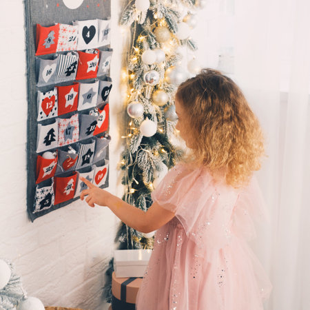 Toddler child gets new advent calendar task, waiting for Christmas. Festive mood for cute curly girl. Kid takes advent bag with dates from the wall at home. From the backの写真素材